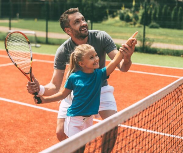 Tennis is fun when father is near. Cheerful father in sports clothing teaching his daughter to play tennis while both standing on tennis court