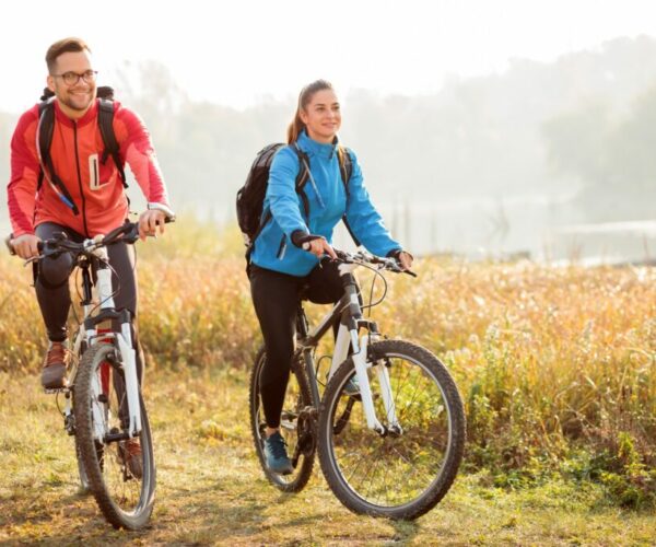 Beautiful happy young couple enjoying early morning bicycle ride by the river or lake. Sunrise through the mist above water in the background